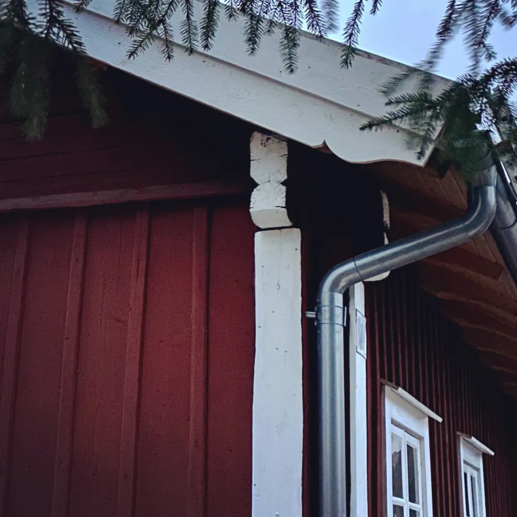Detalj av rött träbyggnads takfot med vit stenpelare, takränna och stuprör av metall